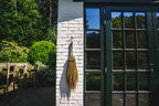Balai mural décoratif en raphia naturel à franges H75 cm contre mur blanc