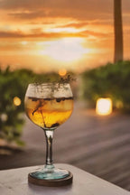 Verre à cocktail avec boisson dorée, herbes et myrtilles sur un dessous de verre rond en racine de teck